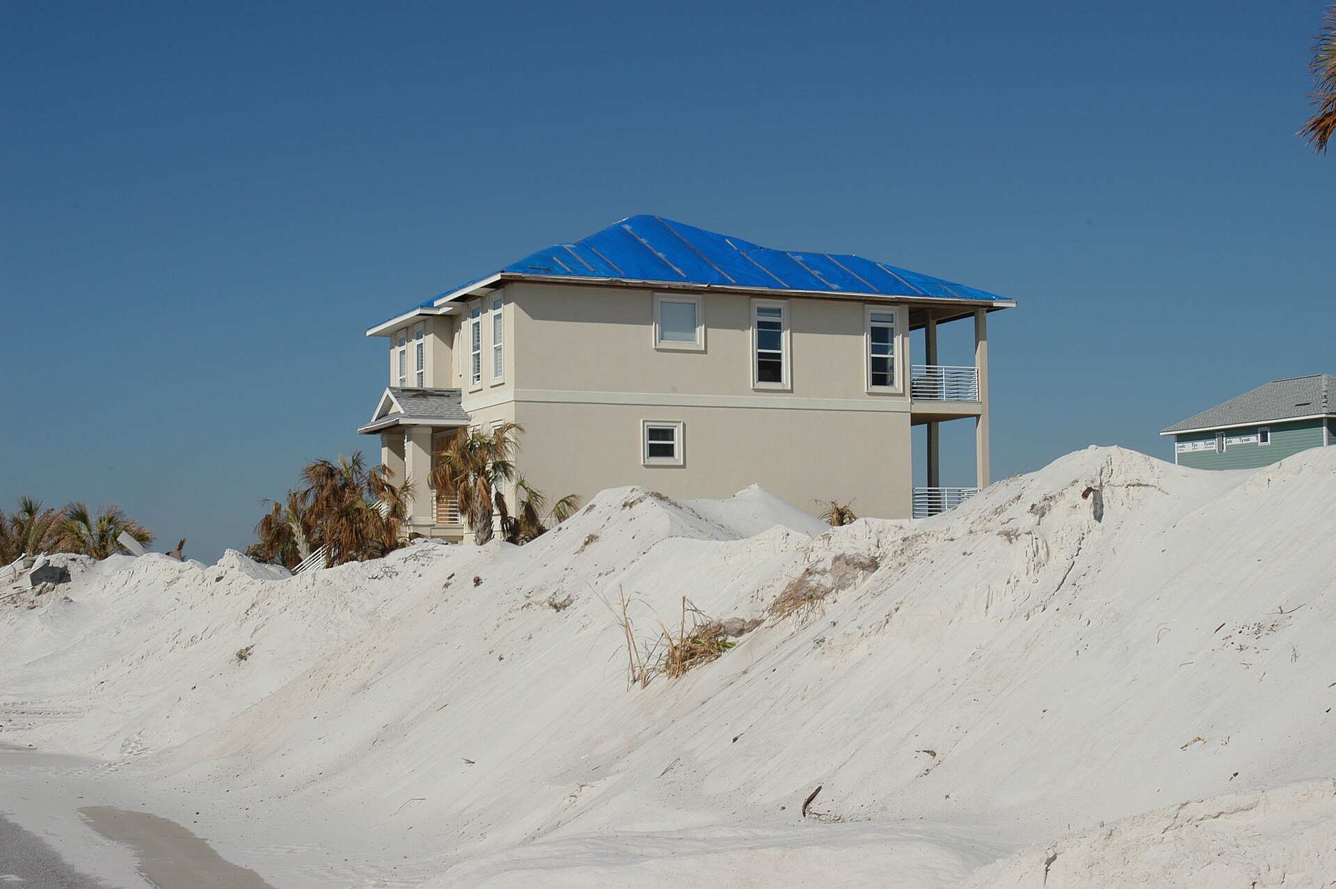 A two-story coastal Florida house with a blue tarp covering its damaged roof, storm-pushed sand banked up against the property